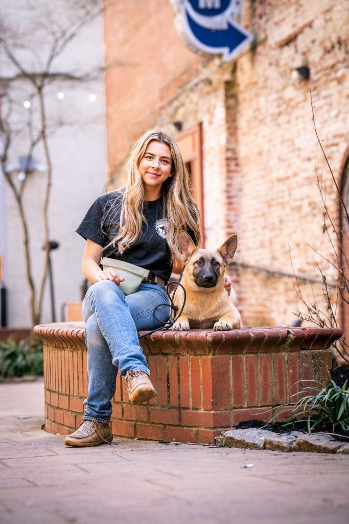Rebeka, dog trainer at K9 Services Unlimited, sitting on a brick planter beside a small dog