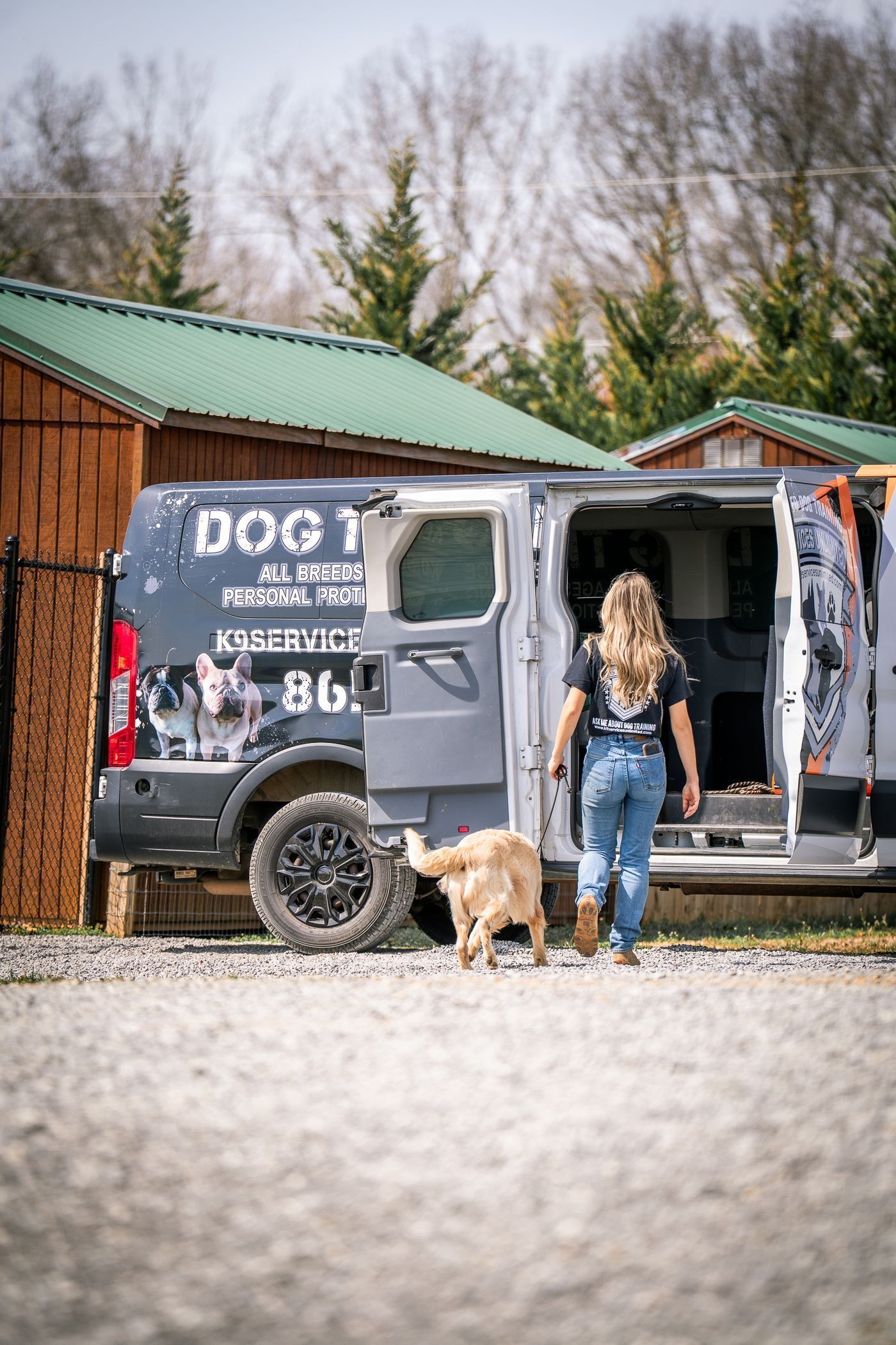 Woman standing with a dog beside a gray van at a campsite with wooden cabins and trees.