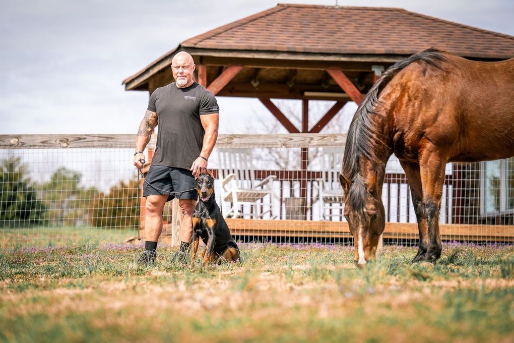 Man standing with a black dog beside a horse in a fenced pasture near a wooden gazebo