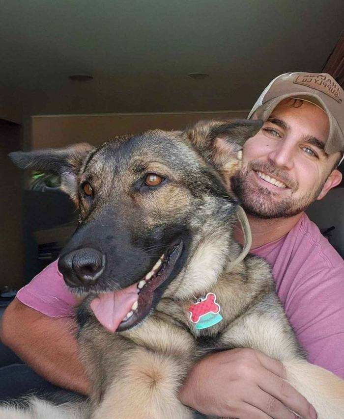 Man holding a happy tan-and-black dog, both smiling indoors