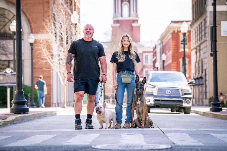 Two people walking dogs on a city street with brick buildings