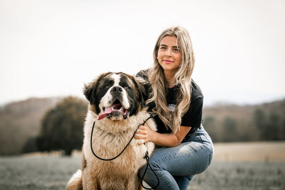 Woman crouches beside a large black-and-white dog in a grassy field, both facing the camera.